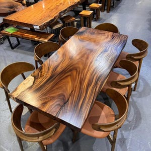 May include: A large rectangular dining table featuring a natural live edge wood slab with prominent dark brown grain patterns, accompanied by six curved wooden chairs with tan upholstered seats arranged around the table in a showroom setting.