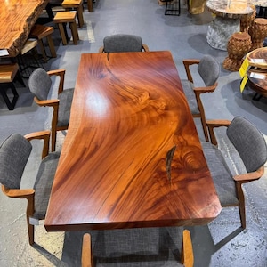 Op de afbeelding: Een rechthoekige eettafel met een blad met natuurlijke randen van gepolijst roodbruin hout met opvallende natuurlijke houtnerven, vergezeld van zes stoelen met houten frames en grijze stoffen bekleding op de zittingen en rugleuningen.