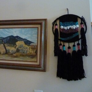 May include: A black and white woven wall hanging with colorful tassels. The hanging features a geometric pattern with pink, blue, and white accents.