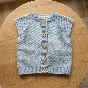May include: A light blue sleeveless knit vest featuring a textured stitch pattern, a ribbed crew neckline, and a ribbed hem. The front opening is secured with five round, light brown wooden buttons arranged in a vertical line down the center.