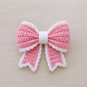 May include: A handmade crochet bow featuring a soft pink center and two long, dangling tails. The entire bow is edged with a delicate, scalloped white border, and a small white band is wrapped tightly around the center to hold the shape together.