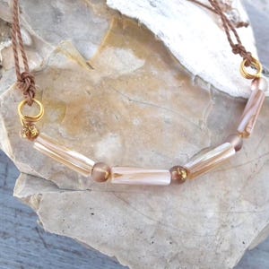 May include: A necklace featuring a brown cord with gold-toned metal rings connecting to a series of cylindrical, pearlescent light pink beads interspersed with smaller, round, metallic-flecked brown beads in a repeating, symmetrical pattern.