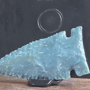 May include: A translucent, light blue arrowhead crafted from stone with a serrated edge and a notched base. The object features a textured, flaked surface and is displayed in a horizontal position, resting on a small black stand against a dark background.