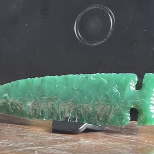 May include: A vibrant green, translucent stone arrowhead featuring a sharp, pointed tip and a notched base. The surface displays a textured, flaked pattern characteristic of knapped stone, and it is supported by a small, black plastic display stand.