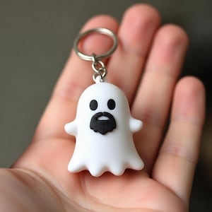 May include: A white ghost keyring featuring a smooth, rounded body with two solid black oval eyes and a small black beard centred on the face. The ghost is attached to a circular silver metal split ring by a short silver metal chain link.