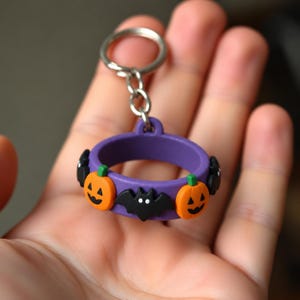 May include: A purple circular keychain featuring alternating orange jack-o'-lanterns with green stems and black bats with white eyes. The charm is attached to a silver-toned metal split ring by a short chain, creating a festive Halloween accessory.
