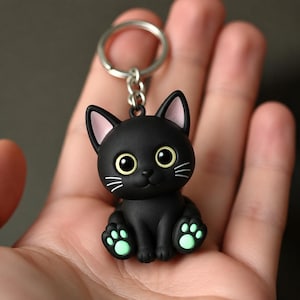 May include: A black cat keychain featuring a matte finish, large yellow eyes, pink inner ears, and light green paw pads. The cat figure is attached to a silver-toned metal split ring by a short chain, creating a compact accessory for keys.