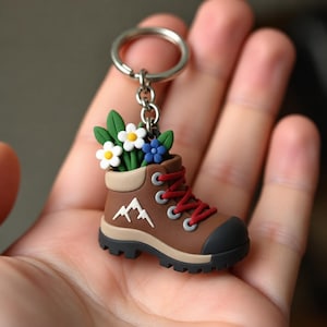 May include: A brown hiking boot keychain featuring a white mountain graphic on the side, red laces, and a black toe cap. The boot serves as a planter for three small flowers with white and blue petals and green leaves attached to a metal ring.