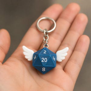 Peut inclure: Un porte-clés en forme de dé à vingt faces bleu avec des chiffres blancs et deux petites ailes blanches fixées sur les côtés. Le dé affiche le chiffre 20 au centre, avec les chiffres 2, 8 et 14 visibles sur les faces triangulaires adjacentes.