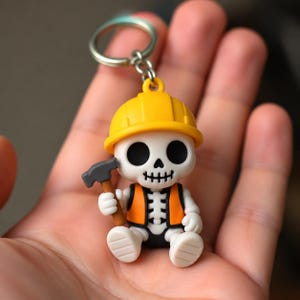 May include: A keychain featuring a white skeleton figurine wearing a bright yellow hard hat and an orange safety vest. The skeleton has large black eye sockets and holds a small gray hammer with a brown handle in its right hand.