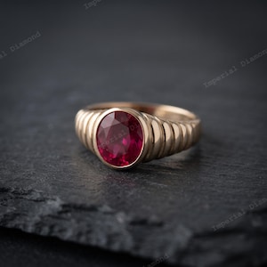Könnte beinhalten: Ein goldener Ring mit einem zentralen, oval geschliffenen, tiefroten Edelstein in einer Zargenfassung. Die Ringschiene ist mit einer Reihe vertikaler, abgerundeter Rillen versehen, die rundherum verlaufen und dem Metall eine texturierte Optik verleihen.