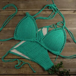 Puede incluir: Un conjunto de bikini de ganchillo en color verde azulado brillante que consta de dos copas triangulares y una braga a juego. La prenda presenta una textura de punto detallada e incluye cordones largos y finos para ajustar en el cuello y los lados.