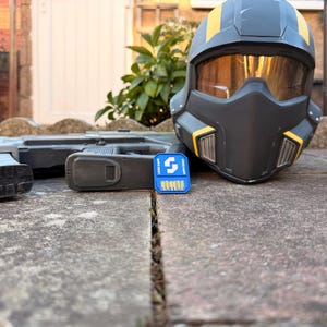 Pode incluir: Um capacete cinza-escuro fosco com detalhes amarelos e uma viseira dourada, posicionado ao lado de um rifle de brinquedo cinza-escuro e um pequeno cartão de memória retangular azul com um logotipo branco e o texto Super Earth impresso.