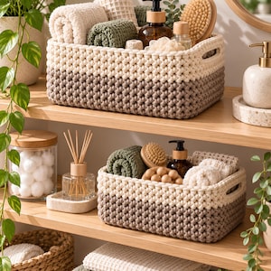 Pode incluir: Dois cestos de armazenamento retangulares feitos de corda trançada grossa em creme e castanho. Os cestos possuem pegas laterais integradas e contêm toalhas texturizadas enroladas em creme e verde-azeitona, escovas de madeira e frascos de vidro.