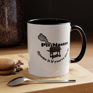 May include: A white ceramic mug with a black interior and handle. The front features a graphic of a charcoal grill with smoke rising, the text Pit Master, and the phrase Smoke it if you've got it printed in black lettering on the white surface.