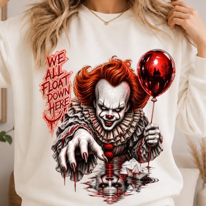 Puede incluir: Una sudadera color crema con un gráfico de un payaso de pelo rojo brillante, cuello con volantes y uñas afiladas. El payaso sostiene un globo rojo y extiende la mano hacia un barco de papel en el agua. El texto dice: WE ALL FLOAT DOWN HERE.