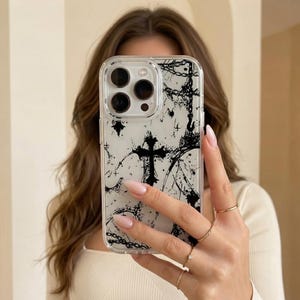 Peut inclure: Une coque de smartphone transparente avec un motif graphique noir représentant une croix, des chaînes et des éclaboussures abstraites. La coque est rectangulaire avec des coins arrondis et une découpe pour un triple capteur photo.