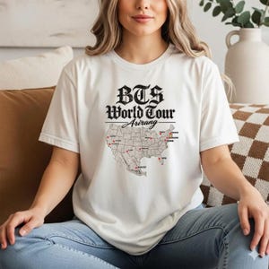 May include: A white short-sleeved t-shirt featuring black text that reads BTS World Tour Arirang above a map of the United States. The map includes red dots marking various cities with corresponding city names printed in black text next to them.