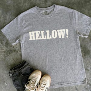May include: A heather grey short-sleeved t-shirt featuring the word HELLOW! printed in large white capital letters across the chest. The shirt is paired with dark grey denim jeans and a pair of white trainers with black stripes.