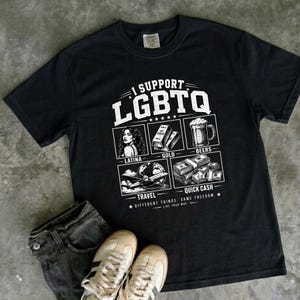 以下が含まれることがあります： I SUPPORT LGBTQと書かれた白いグラフィックテキストが特徴の黒い半袖Tシャツ。テキストの下には、LATINA、GOLD、BEERS、TRAVEL、QUICK CASHとラベル付けされた4つの正方形のイラストがあり、DIFFERENT THINGS, SAME FREEDOMというフレーズが添えられています。
