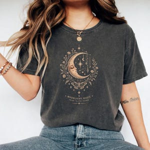 May include: A charcoal grey short-sleeve t-shirt featuring a gold-toned graphic of a crescent moon with a face, surrounded by floral vines, stars, and a moth. The text "MOONLIGHT MAGIC" and "DREAM WANDER WONDER" is printed below the design.