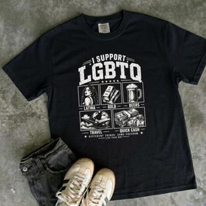 May include: A black short-sleeve t-shirt featuring a white graphic print that reads I SUPPORT LGBTQ. The design includes illustrations of a person, gold bars, a beer mug, an airplane, and stacks of cash with labels Latina, Gold, Beers, Travel, and Quick Cash.