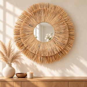 May include: A round wall mirror featuring a double-layered frame made of natural tan raffia or straw fibers arranged in a sunburst pattern. The central circular glass mirror reflects a bright interior space with white flowers.