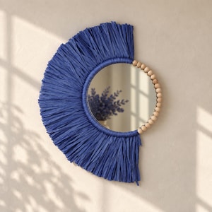 May include: A circular wall mirror featuring a decorative frame partially adorned with deep blue raffia fringe and a section of natural light-colored wooden beads. The mirror surface reflects a blurred view of a purple floral arrangement.