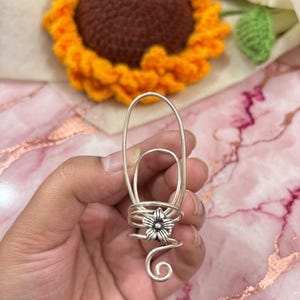 May include: A silver-toned metal hair accessory featuring a large oval loop at the top, a coiled wire base, and a small five-petaled flower charm centered on the coil. The wire design ends in a decorative spiral curl at the bottom of the piece.