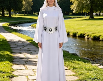 Puede incluir: Un vestido blanco largo hasta el suelo con capucha, mangas largas y anchas, y cuello alto. La prenda presenta un cinturón plateado metálico con detalles rectangulares negros y un adorno geométrico central, logrando un aspecto estructurado.
