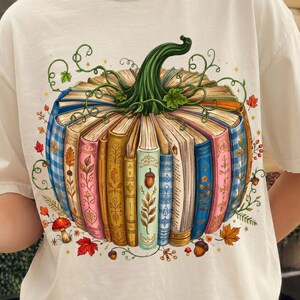 May include: A cream-colored t-shirt featuring a graphic print of a pumpkin constructed from a circular arrangement of colorful, vintage-style books. The design includes green vines, small mushrooms, and autumn leaves surrounding the book pumpkin.