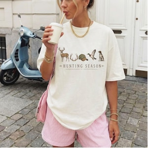 May include: An off-white short-sleeve t-shirt featuring a graphic print of a deer, antlers, a baseball cap, a duck, and a dog, with the text "HUNTING SEASON" printed below. The shirt is paired with light pink striped shorts and a gold chain necklace.