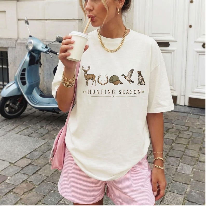 May include: An off-white short-sleeve t-shirt featuring a graphic print of a deer, antlers, a baseball cap, a duck, and a dog, with the text "HUNTING SEASON" printed below. The shirt is paired with light pink striped shorts and a gold chain necklace.