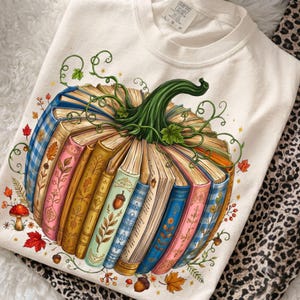 May include: A cream-colored t-shirt featuring a graphic of a pumpkin constructed from stacked, colorful vintage books. The pumpkin has a green stem and vines, surrounded by small illustrations of red mushrooms, autumn leaves, and acorns.