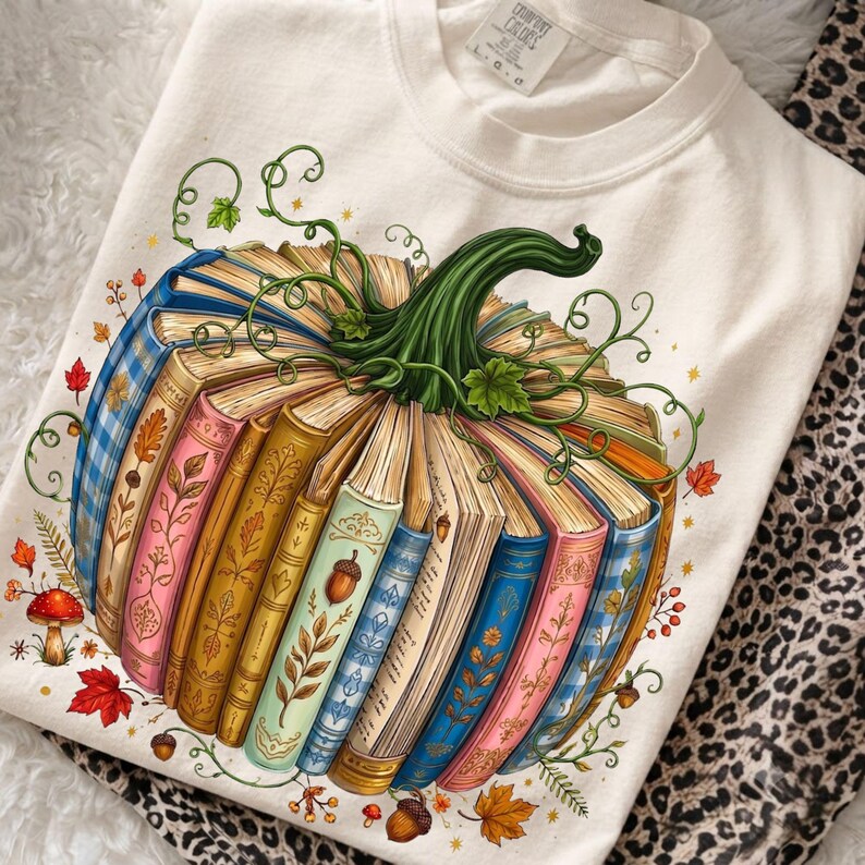 May include: A cream-colored t-shirt featuring a graphic of a pumpkin constructed from stacked, colorful vintage books. The pumpkin has a green stem and vines, surrounded by small illustrations of red mushrooms, autumn leaves, and acorns.