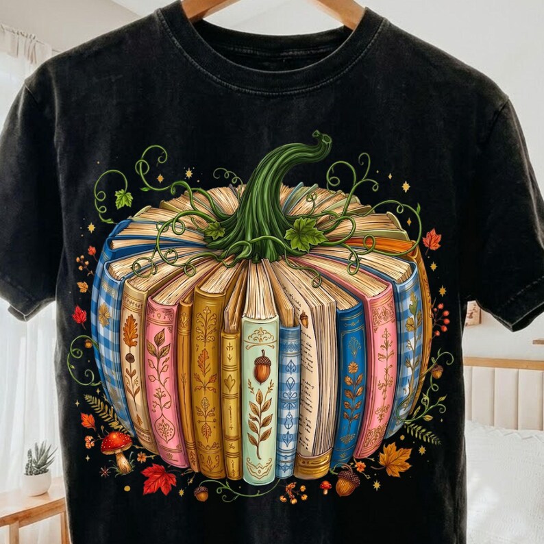 May include: A black t-shirt featuring a graphic of a pumpkin constructed from a circular arrangement of colorful books. The pumpkin has a green stem and vines, surrounded by small illustrations of autumn leaves, acorns, and mushrooms.