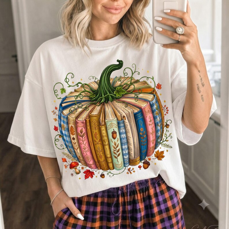 May include: A white t-shirt featuring a graphic of a pumpkin constructed from a collection of colorful, upright books. The pumpkin has a green stem and vine accents, surrounded by small illustrations of autumn leaves, acorns, and mushrooms.