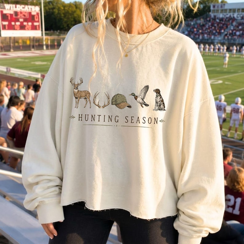 May include: A cream-colored crewneck sweatshirt featuring a graphic print with a deer, antlers, a baseball cap, a flying duck, and a dog. The text "HUNTING SEASON" is printed in black below the illustrations, centered on the front of the garment.