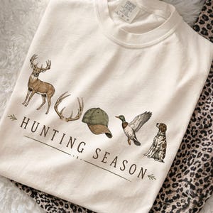 May include: A cream-colored t-shirt featuring a graphic print of a deer, antlers, a green baseball cap, a mallard duck, and a spotted dog. The text "HUNTING SEASON" is printed in brown lettering below the illustrations with decorative arrows.