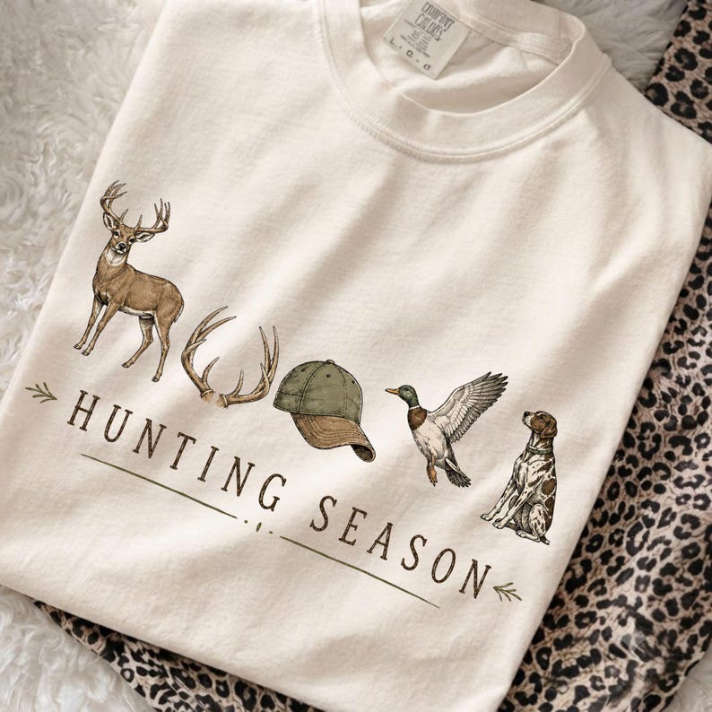 May include: A cream-colored t-shirt featuring a graphic print of a deer, antlers, a green baseball cap, a mallard duck, and a spotted dog. The text "HUNTING SEASON" is printed in brown lettering below the illustrations with decorative arrows.