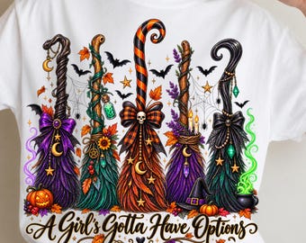 May include: A white t-shirt featuring a graphic of five stylized witch brooms decorated with bows, pumpkins, stars, moons, and crystals. The text "A Girl's Gotta Have Options" is printed in a black cursive font at the bottom with autumn leaves.