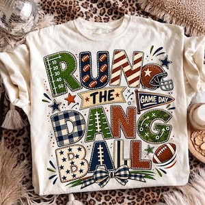 May include: A cream-colored t-shirt featuring the text RUN THE DANG BALL in large, patterned letters. The design includes football-themed graphics like a field grid, footballs, a helmet, a pennant flag, and a blue and white gingham bow.
