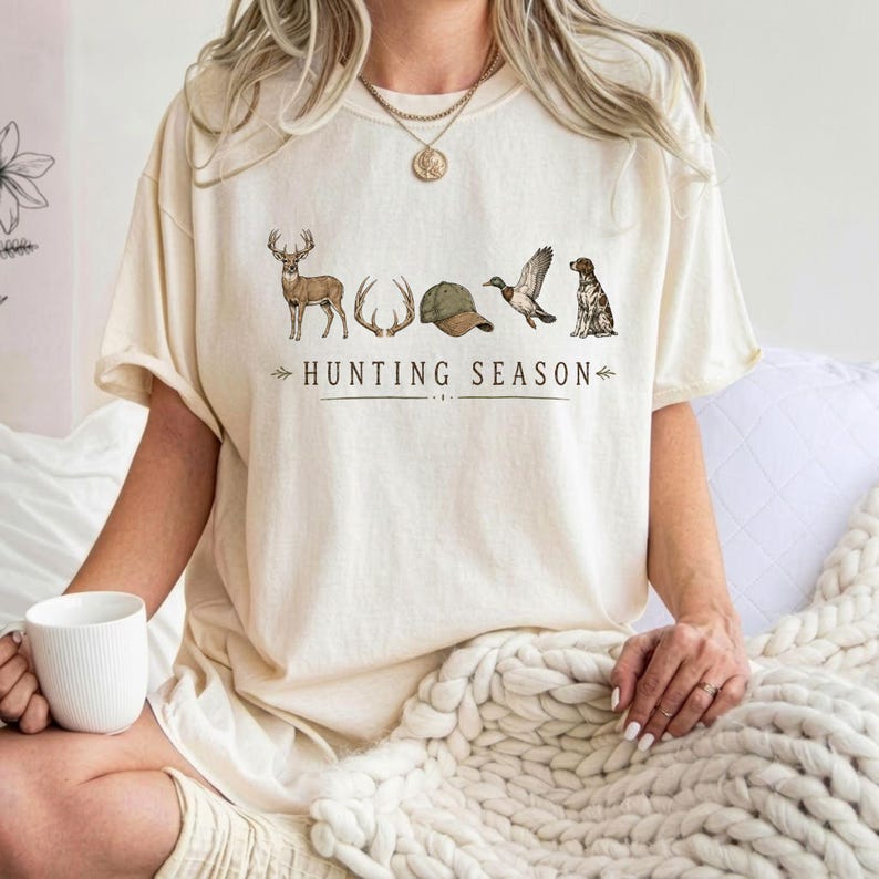 May include: A cream-colored t-shirt featuring a graphic print of a deer, antlers, a green cap, a flying duck, and a dog. The text "HUNTING SEASON" is printed in black capital letters below the illustrations. The shirt has short sleeves and a crew neck.
