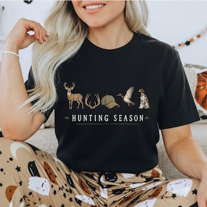 May include: A black short-sleeved t-shirt featuring a graphic print with a deer, antlers, a baseball cap, a flying duck, and a hunting dog. Below the illustrations, the text reads "HUNTING SEASON" in white capital letters with decorative lines.