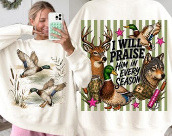 May include: A cream-colored crewneck sweatshirt featuring a graphic print of two mallard ducks in flight and among reeds on the front. The back displays a deer, a wolf, and a mallard duck with the text I WILL PRAISE HIM IN EVERY SEASON.