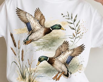 May include: A white short-sleeve t-shirt featuring a watercolor-style graphic of two mallard ducks in flight. The design includes brown and green plumage, blue wing patches, orange feet, and tall brown cattails against a soft green background.
