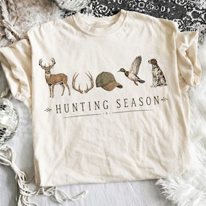 May include: A cream-colored short-sleeve t-shirt featuring a graphic print of a deer, antlers, a green baseball cap, a flying duck, and a sitting dog. Below the illustrations, the text reads HUNTING SEASON in a dark brown serif font.