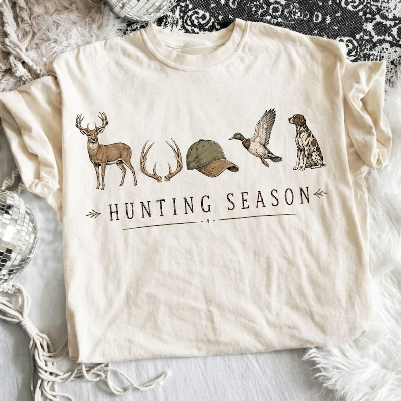 May include: A cream-colored short-sleeve t-shirt featuring a graphic print of a deer, antlers, a green baseball cap, a flying duck, and a sitting dog. Below the illustrations, the text reads HUNTING SEASON in a dark brown serif font.