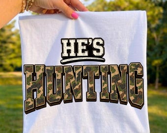 May include: A white t-shirt featuring the text HE'S in black with a white outline above the word HUNTING, which is rendered in a blocky, three-dimensional font filled with a green, brown, and black camouflage pattern and a dark drop shadow.