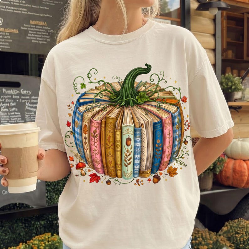 May include: A cream-colored t-shirt featuring a graphic print of a pumpkin constructed from a circular arrangement of colorful, vintage-style books. The design includes green vines, small mushrooms, and autumn leaves surrounding the book pumpkin.